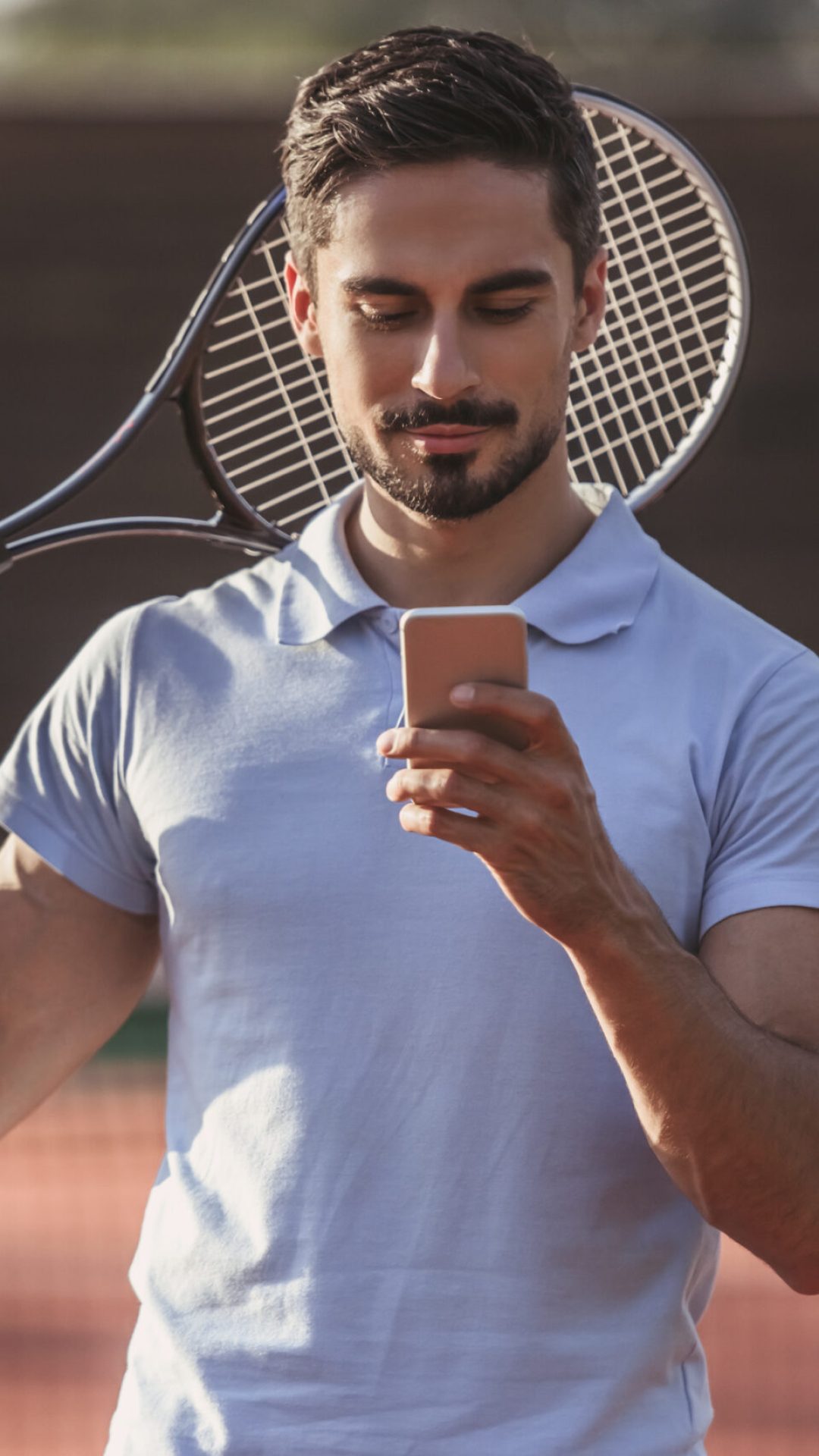Handsome man is using a smart phone while playing tennis on tennis court outdoors